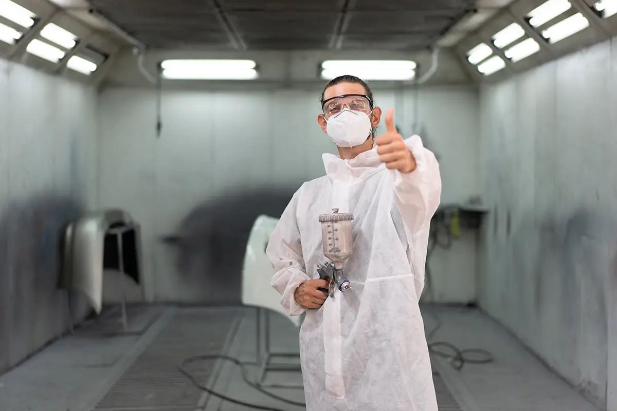 Smiling auto body specialist giving a thumbs-up before painting a vehicle.